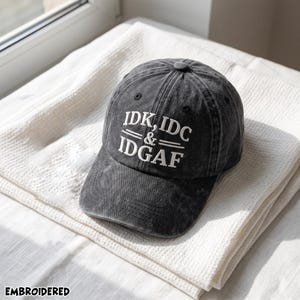 May include: A charcoal gray, acid-washed baseball cap featuring white embroidered text on the front that reads IDK, IDC & IDGAF. The cap has a structured crown, a curved brim with visible stitching, and a button at the top center of the crown.