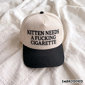 Może przedstawiać: Kremowa czapka z daszkiem z czarnym daszkiem i czarnym guzikiem na górze. Na przednim panelu znajduje się czarny haftowany napis KITTEN NEEDS A FUCKING CIGARETTE. Słowo EMBROIDERED jest nadrukowane w prawym dolnym rogu.