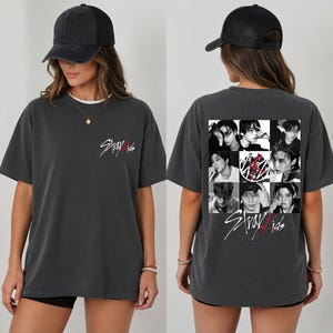 May include: A charcoal gray short-sleeve t-shirt featuring a small white Stray Kids logo on the front left chest. The back displays a grid of eight black and white portraits of the band members, a circular logo, and the Stray Kids text logo.