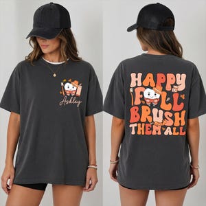 May include: A charcoal gray t-shirt featuring a small graphic on the front left chest of a cartoon tooth wearing a hat and scarf with the name "Askley" below it. The back displays a large graphic with the text "HAPPY FULL BRUSH THEM ALL".