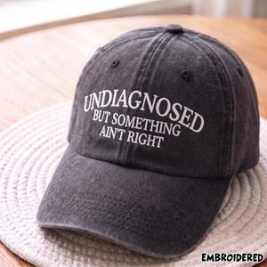 May include: A charcoal grey baseball cap with a distressed, washed-out finish. The front features white embroidered text that reads: UNDIAGNOSED BUT SOMETHING AIN'T RIGHT. The cap has a structured crown, a curved brim, and matching stitching details.