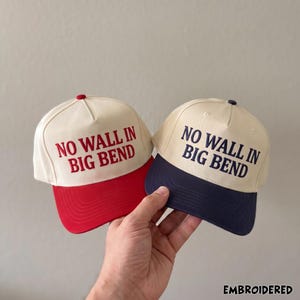May include: Two baseball caps with off-white crowns and embroidered text reading NO WALL IN BIG BEND. One cap features a red brim and red lettering, while the other cap features a navy blue brim and navy blue lettering. Both caps have a button top.
