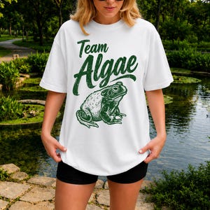 May include: A white short-sleeve t-shirt featuring a dark green graphic print of a toad and the text "Team Algae" in a distressed, stylized font. The design is centered on the chest, showcasing a detailed illustration of a toad in profile.
