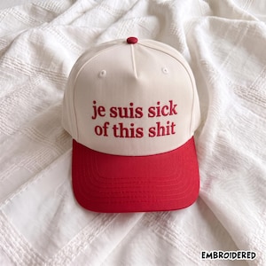 Peut inclure: Une casquette de baseball crème avec une visière rouge et le texte brodé en rouge sur le devant qui indique "je suis sick of this shit". La casquette présente une calotte structurée avec un petit bouton rouge au sommet et des coutures visibles.