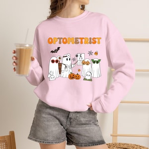 Könnte beinhalten: Ein hellrosa Langarm-Sweatshirt mit dem orangefarbenen Schriftzug OPTOMETRIST und einer Grafik von Geistern in einer Arztpraxis. Das Design zeigt einen Geist mit Stirnspiegel, eine Sehtafel, Kürbisse, eine Fledermaus und Zuckermais.