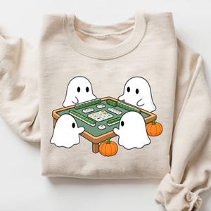 May include: Beige sweatshirt featuring a cartoon illustration of four white ghosts playing mahjong around a green table. Two orange pumpkins are placed near the table. The sweatshirt has a crew neck and long sleeves.