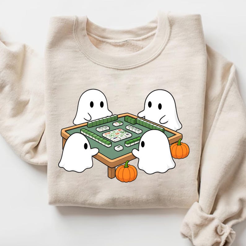 May include: Beige sweatshirt featuring a cartoon illustration of four white ghosts playing mahjong around a green table. Two orange pumpkins are placed near the table. The sweatshirt has a crew neck and long sleeves.