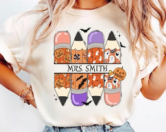 Könnte beinhalten: Ein cremefarbenes T-Shirt mit einer Grafik von acht Bleistiften in zwei Reihen. Die Bleistifte sind mit Mustern aus Kürbissen, Geistern, Fledermäusen und Zuckermais verziert. Der Text "MRS. SMITH" steht in Schwarz in der Mitte.