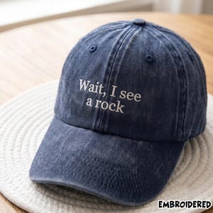 May include: A dark blue baseball cap with a distressed, washed-out finish. The front of the cap features white embroidered text that reads, Wait, I see a rock. The cap has a structured crown, a curved brim, and stitched eyelets for ventilation.