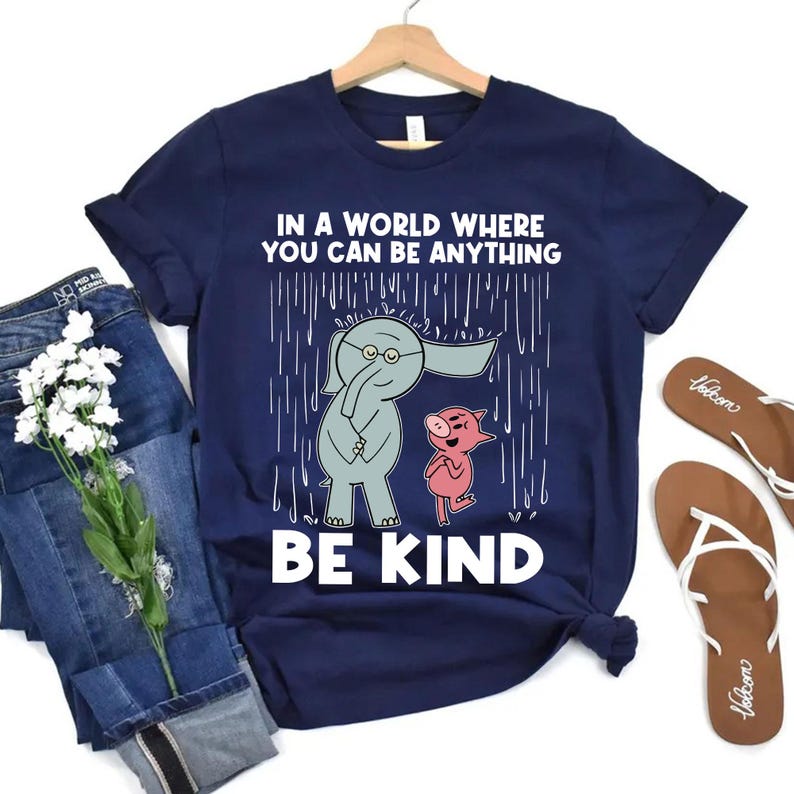 May include: A navy blue short-sleeve t-shirt featuring a graphic of a grey elephant and a pink pig standing in the rain. The text reads, "IN A WORLD WHERE YOU CAN BE ANYTHING BE KIND." The shirt is displayed with blue jeans and brown flip-flops.