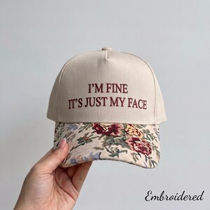 May include: A beige baseball cap featuring the embroidered text I'M FINE IT'S JUST MY FACE in dark red lettering. The brim of the cap is decorated with a multi-coloured floral pattern in shades of red, yellow, and blue on a beige background.