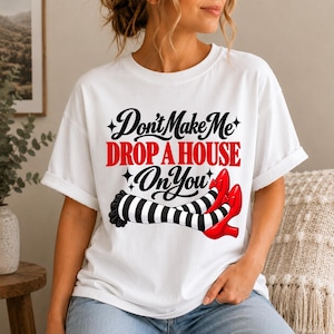 Könnte beinhalten: Ein weißes Kurzarm-T-Shirt mit dem schwarzen und roten Text "Don't Make Me DROP A HOUSE On You" über einer Grafik von zwei Beinen in schwarz-weiß gestreiften Strümpfen und leuchtend roten Schuhen mit Schleifen, akzentuiert durch kleine schwarze Sterne.