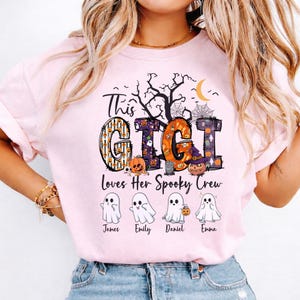 May include: A light pink t-shirt featuring a graphic with the text "This GIGI Loves Her Spooky Crew". The design includes large letters filled with Halloween patterns, a crescent moon, a tree, and four ghost illustrations labeled James, Emily, Daniel, and Emma.