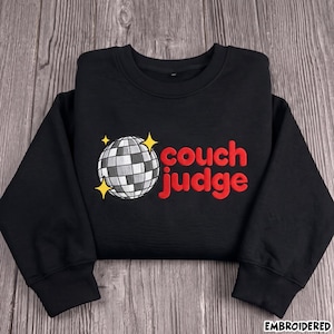 May include: A black long-sleeve sweatshirt featuring an embroidered graphic of a silver disco ball with yellow star accents. To the right of the disco ball, the text "couch judge" is embroidered in bold red lowercase letters on the chest.