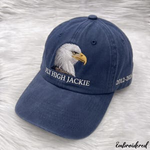 May include: A navy blue baseball cap featuring an embroidered bald eagle head on the front panel. Below the eagle, the text reads FLY HIGH JACKIE in white. The side of the cap displays the embroidered white text 2012-2026 on the right side.