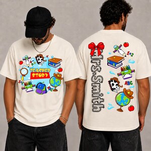 May include: A cream-colored t-shirt featuring a school-themed graphic. The front displays the text "TEACHER STORY" surrounded by illustrations of a globe, books, a backpack, and crayons. The back features a red bow and the text "Mrs. Smith".