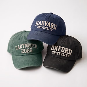 May include: Three baseball caps in navy blue, dark green, and black. The navy cap features the text HARVARD UNIVERSITY, the green cap displays DARTMOUTH 2026, and the black cap shows OXFORD UNIVERSITY. Each cap has a curved brim and embroidered lettering.
