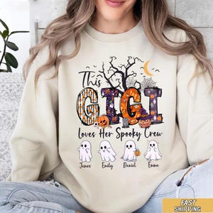 May include: A cream-colored crewneck sweatshirt featuring a graphic with the text This GIGI Loves Her Spooky Crew. The design includes a black tree, spiderwebs, a crescent moon, and four white ghost illustrations labeled James, Emily, Daniel, and Emma.