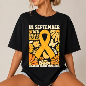 May include: A black t-shirt featuring a large yellow awareness ribbon in the center, surrounded by orange and yellow floral and butterfly graphics. The text reads: IN SEPTEMBER WE WEAR GOLD CHILDHOOD CANCER AWARENESS in a bold, retro-style font.