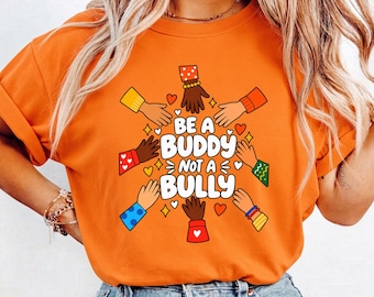 Puede incluir: Una camiseta naranja de manga corta con un gráfico de varias manos de diferentes tonos de piel que apuntan hacia el centro. El texto dice BE A BUDDY NOT A BULLY, rodeado de pequeños iconos de corazones y estrellas en rojo, amarillo y blanco.