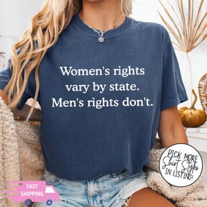 May include: A navy blue short-sleeve t-shirt featuring white text that reads: Women's rights vary by state. Men's rights don't. The shirt has a crew neckline and a relaxed fit, showcasing a bold, centered typographic design on the front chest area.