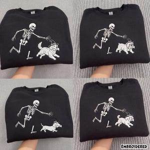 May include: A black crewneck sweatshirt featuring an embroidered white skeleton reaching towards a dog wearing a witch hat and holding a bone. Four variations are shown, each depicting a different dog breed: a golden retriever, bulldog, dachshund, and poodle.