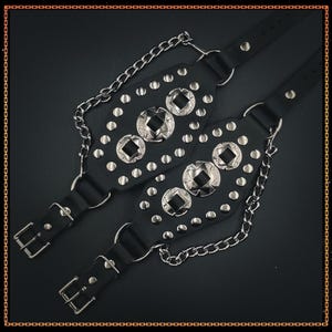 Puede incluir: Dos pulseras de cuero negro con tachuelas metálicas plateadas y adornos cuadrados. Cada pulsera tiene un cierre de hebilla y está conectada por una cadena plateada. Fondo oscuro.