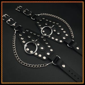 Könnte beinhalten: Paar schwarze Leder-Handgelenksfesseln mit silberfarbenen Metallnieten, Ringen und Kettenakzenten. Jede Fessel verfügt über verstellbare Schnallenverschlüsse. Das Design ist symmetrisch und detailliert.