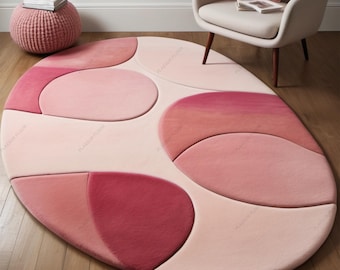 May include: An oval-shaped rug in shades of pink and rose. The rug features a geometric design with large, rounded shapes in varying tones, creating a soft, textured appearance. The rug is placed on a wooden floor, with a chair and ottoman in the background.