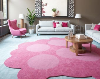 May include: A pink and white cloud-shaped rug with a textured surface. The rug has five circles in the center, with the largest circle in the middle. The rug is in a living room with a gray sofa, a pink armchair, and a wooden coffee table.