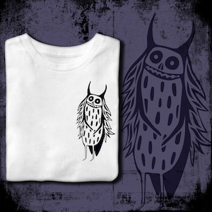 Peut inclure: Un t-shirt blanc avec une illustration noire et blanche d'un monstre avec des cornes, un sourire narquois et un corps velu. Le monstre est debout, les bras croisés.