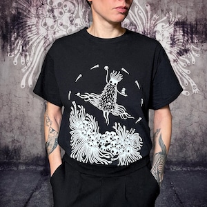 Könnte beinhalten: Schwarzes T-Shirt mit einem weißen Grafikdesign einer Kreatur mit langen, fließenden Ranken und Totenköpfen. Das Design befindet sich auf der Vorderseite. Das Shirt hat kurze Ärmel und einen Rundhalsausschnitt.