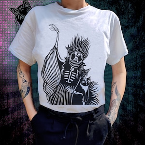 Könnte beinhalten: Weißes T-Shirt mit einem schwarz-weißen Grafikdruck eines Skeletts mit langen Haaren und zwei Katzen. Das Skelett trägt ein Kleid und hält eine der Katzen.