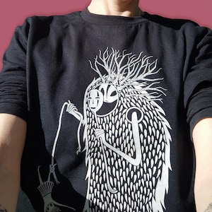 Könnte beinhalten: Schwarzes Langarmshirt mit einem weißen Grafikdruck eines Wesens mit einer Gesichtsmaske und einem baumartigen Kopf. Das Wesen hält eine Schnur, an der eine kleine Figur befestigt ist.