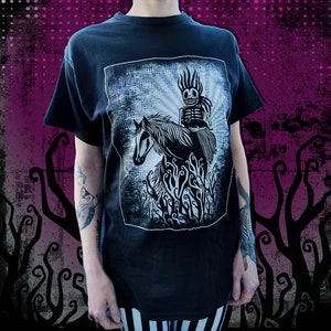 Könnte beinhalten: Schwarzes T-Shirt mit einer weiß-grauen Grafik eines Skeletts auf einem Pferd. Das Design hat einen verwitterten, strukturierten Hintergrund. Das Skelett hat einen stacheligen Kopf und sitzt auf dem Pferd. Das Pferd ist von stilisiertem, wirbelndem Laub umgeben.