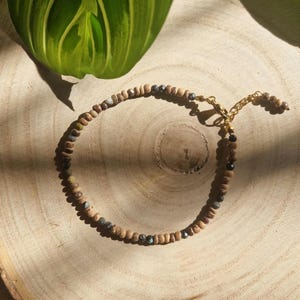 Könnte beinhalten: Ein braunes und schwarzes Perlenarmband mit einem goldenen Verschluss. Das Armband besteht aus kleinen Holzperlen und schwarzen Edelsteinperlen.