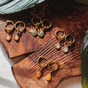 Könnte beinhalten: Goldfarbene Creolen mit tropfenförmigen Edelsteinanhängern in verschiedenen Farben, darunter Pfirsich, Grün und Weiß. Die Ohrringe werden auf einer Holzoberfläche präsentiert. Schmuck.