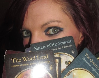 May include: A collection of rectangular oracle cards featuring dark borders and gold text. The cards display titles such as The Word Lord, Sisters of the Seasons, and The Queen of, accompanied by illustrations of figures and symbolic imagery.