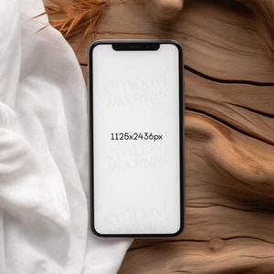 Blank black smartphone on natural wood with soft white fabric and dried leaves, creating a warm, organic setting for mobile design presentations