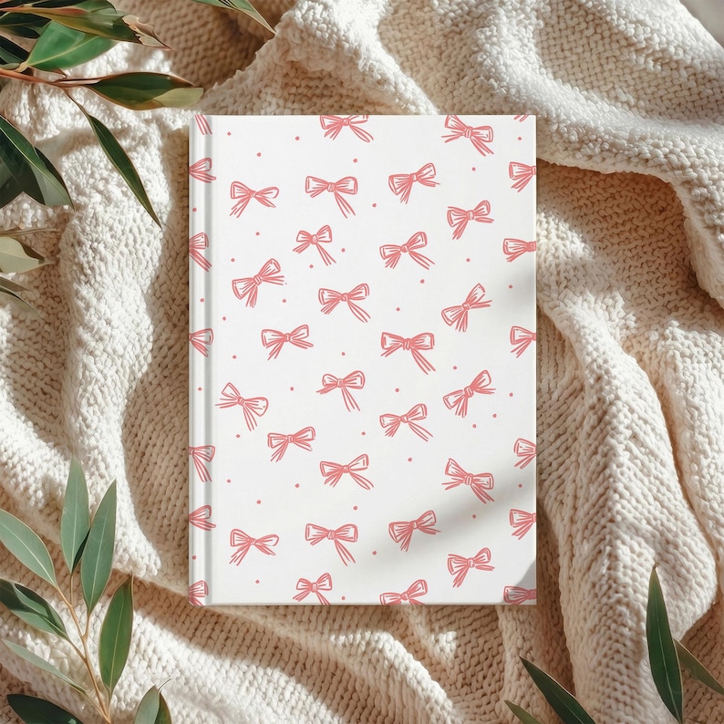 May include: A white journal with a pink bow pattern. The bows are evenly spaced across the cover, with small pink dots between them. The journal is resting on a cream-colored knitted blanket, with green leaves in the corners.