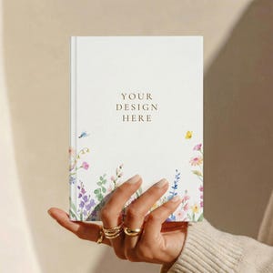 May include: A white hardback notebook featuring a watercolour illustration of colourful wildflowers and a small yellow butterfly at the bottom. The centre of the cover displays the text YOUR DESIGN HERE in a simple, dark brown serif font.