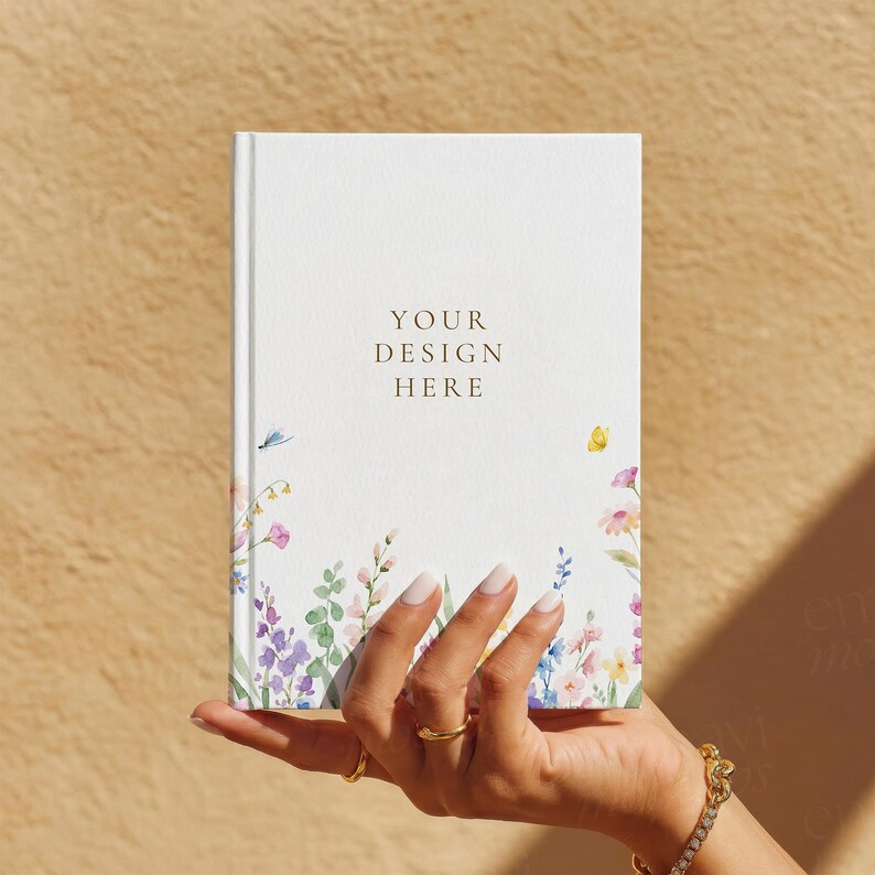 May include: A white hardback journal featuring a watercolour floral border at the bottom with various colourful wildflowers and a small yellow butterfly. The centre of the cover displays the gold-coloured text YOUR DESIGN HERE in a serif font.