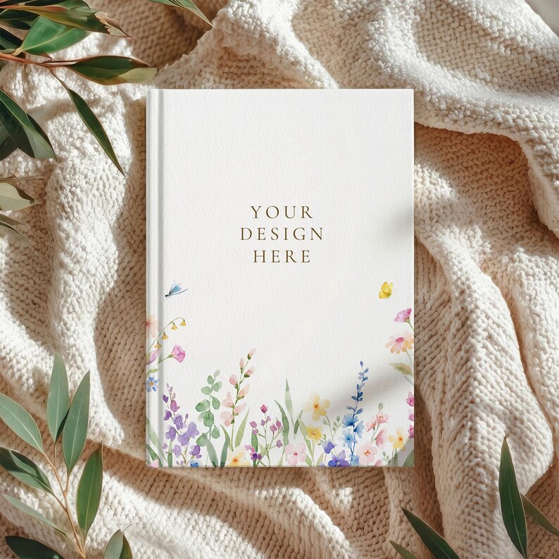 May include: A white journal with the text "YOUR DESIGN HERE" in a serif font. The bottom edge features a watercolor floral design with pink, purple, yellow, and blue flowers. The journal is resting on a cream-colored knitted blanket.