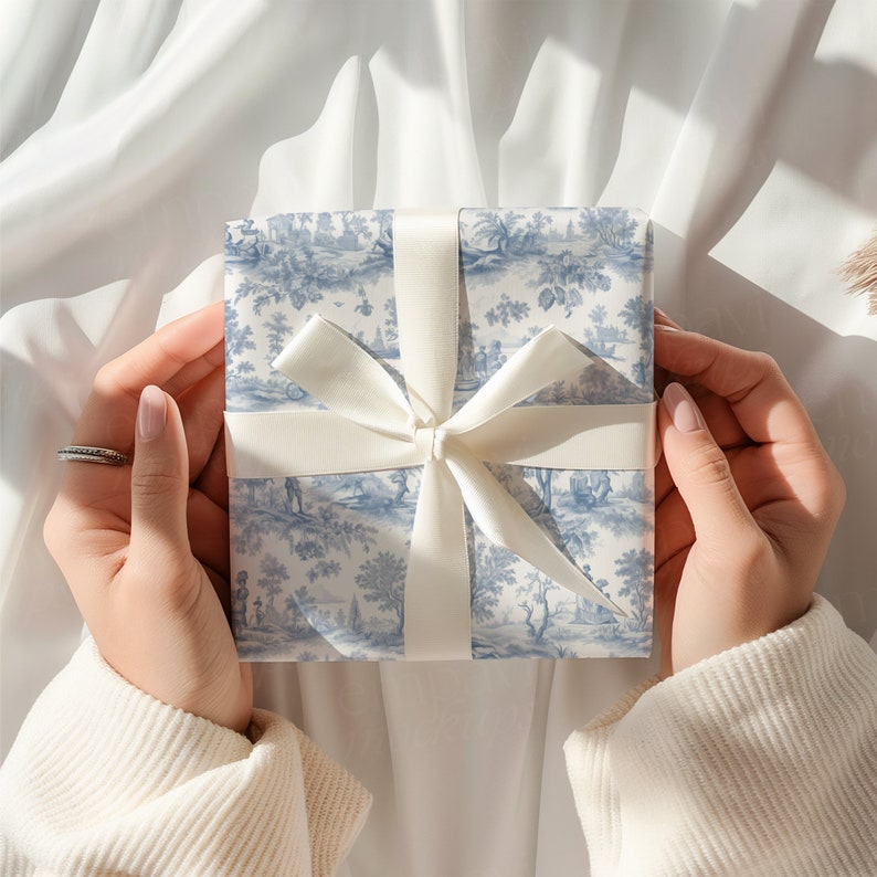 K&ouml;nnte beinhalten: Eine quadratische Geschenkbox, eingewickelt in blau-wei&szlig;es Toile-de-Jouy-Papier mit detaillierten pastoralen Szenen aus B&auml;umen und Figuren. Die Box ist mit einem glatten, cremefarbenen Satinband in einem zentrierten Kreuzmuster verschn&uuml;rt.