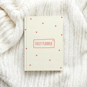 May include: A cream-colored daily planner with a hard cover featuring small red heart illustrations scattered across the front. The center of the cover displays the text DAILY PLANNER enclosed within a simple red rectangular outline.