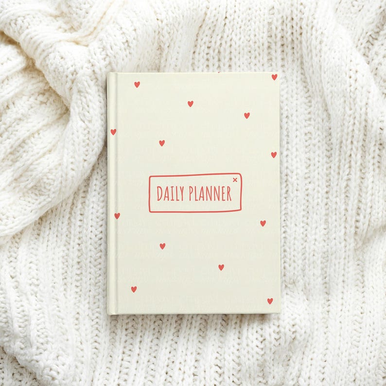 May include: A cream-colored daily planner with a hard cover featuring small red heart illustrations scattered across the front. The center of the cover displays the text DAILY PLANNER enclosed within a simple red rectangular outline.