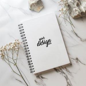 May include: A white spiral bound notebook with the text "your design here" on the cover. The notebook is on a white marble surface with white flowers and a rock.