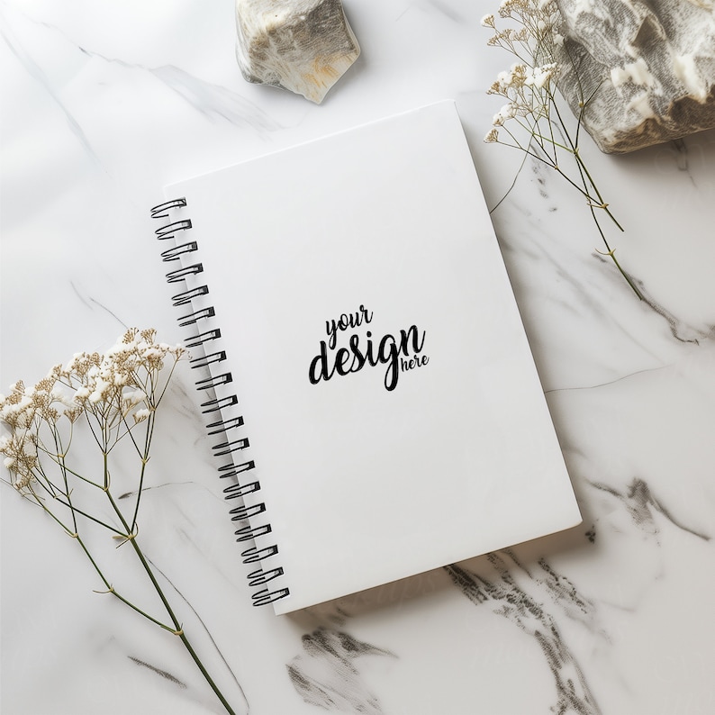 May include: A white spiral bound notebook with the text "your design here" on the cover. The notebook is on a white marble surface with white flowers and a rock.