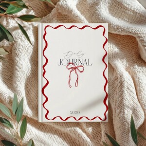 May include: A white hardcover daily journal featuring a red wavy border, a red line-art bow illustration, and black text that reads Daily JOURNAL 2030. The journal has a clean, centered layout with elegant typography and a minimalist aesthetic.