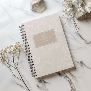 May include: A beige daily planner with a white floral pattern and the text "daily planner 2025" on the cover. The planner is lying on a white marble surface with white flowers.